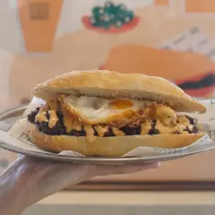 Un delicioso almuerzo valenciano: un bocadillo con carne, huevo frito y salsa cremosa, servido en plato metálico.