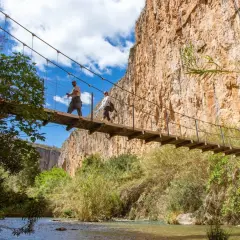 Dos excursionistas cruzan un puente colgante sobre un río, explorando las rutas de senderismo de Valencia.
