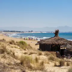 Chiringuito de paja en la playa del Rebollo, con gente disfrutando del sol y el mar azul. Montañas al fondo.