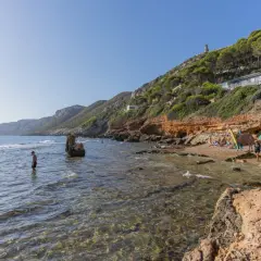 Gente disfruta del sol y el mar en una cala rocosa. Se ven casas y una torre en la **punta negra** de la colina verde.