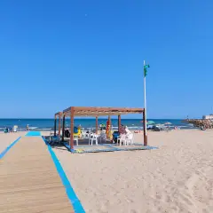 Pasarela de madera azul en la playa de Massamagrell, con chiringuito y gente disfrutando del sol y el mar.