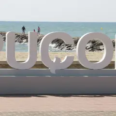 Letras gigantes "PUZOL" en la playa, con el mar azul y personas al fondo.
