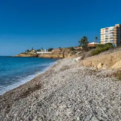 La cala Amerador, con su playa de guijarros y aguas turquesas, se extiende bajo un cielo azul. Edificios modernos y vegetación mediterránea bordean la costa.