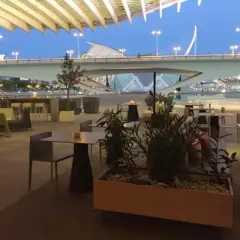 Mesas y sillas en la terraza de restaurantes con vistas a un puente moderno y un cielo azul crepuscular.