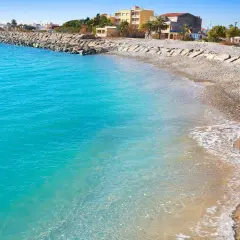 Aguas turquesas bañan la playa almenara, con un rompeolas de rocas y edificios al fondo.