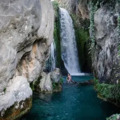 Una mujer se baña en las **fonts de Algar**, rodeada de rocas y vegetación, bajo una cascada de agua cristalina.