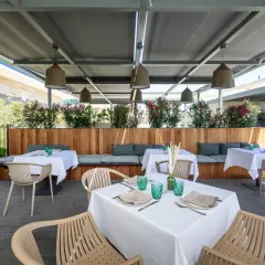 Terraza de la colonia de carmela con mesas puestas, asientos de madera y plantas floridas.