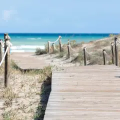 Pasarela de madera que conduce a la playa Raco de la Mar. El mar azul y las dunas de arena crean un paisaje sereno.