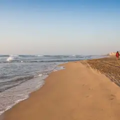 Dos personas pasean por la **playa Babilonia** al atardecer, con el mar y las casas de fondo.