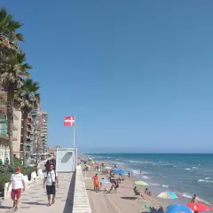 Gente disfrutando del sol y el mar en la playa Mareny de Barraquetes, con palmeras y edificios de apartamentos.