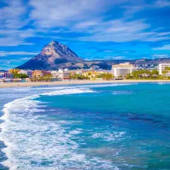 Olas turquesas rompen en la arena dorada de la playa del Arenal Javea, con edificios y el imponente Montgó al fondo.