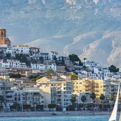 Vista del **castell de l'Olla** coronando la colina, con sus casas blancas y la iglesia de cúpula azul. Un velero navega frente a la costa.