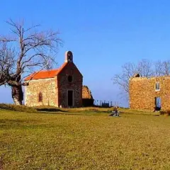 La ermita de Santa Catalina, una pequeña iglesia de piedra con tejado naranja, se alza junto a unas ruinas bajo un cielo azul. Un árbol desnudo y un paisaje verde completan la escena.