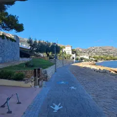 Paseo marítimo con siluetas de personas en la cala del Soio. El mar azul y las montañas de fondo crean un paisaje idílico.