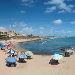 Gente disfrutando del sol y el mar en la playa Cabo Cervera, con sombrillas de colores y edificios al fondo.