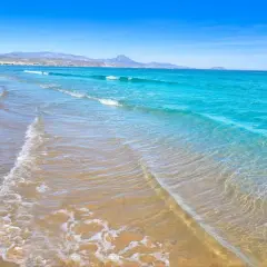 Aguas turquesas y arena dorada en las calas de San Juan, Alicante. El mar Mediterráneo acaricia la costa, con montañas al fondo.