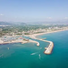 Vista aérea del puerto de Oliva con barcos navegando y la playa llena de gente.