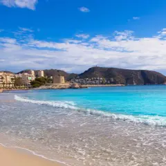 La playa de l'Ampolla en Moraira, con su arena dorada y aguas turquesas, invita a un día de sol y mar. Al fondo, edificios y una torre defensiva se integran en el paisaje.
