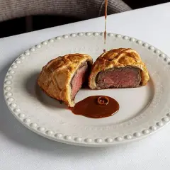 Un Wellington de ternera jugoso, cortado por la mitad, servido con salsa en un plato elegante. Una delicia para probar en los restaurantes de Valencia.