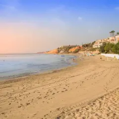 Playa con arena dorada y el mar azul. Al fondo, un barranco rubio con edificios y vegetación.