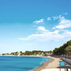 La playa de la Marineta Casiana, con su arena dorada y aguas turquesas, invita a un paseo tranquilo bajo un cielo azul salpicado de nubes. Palmeras y pinos bordean el paseo marítimo.