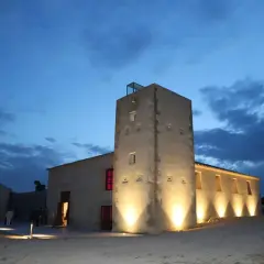La torre Ansaldo, iluminada al anochecer, se alza junto a edificios bajos bajo un cielo azul oscuro con nubes.