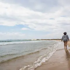 Una persona pasea con su perro por la orilla de la playa de Pinedo, con el mar y el cielo nublado de fondo.
