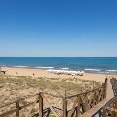 Pasarela de madera hacia la playa Moncayo, con arena dorada, mar azul y sombrillas blancas alineadas.