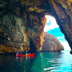 Dos personas en kayak rojo exploran la cueva de la cala del llop mari, con aguas turquesas y formaciones rocosas impresionantes.