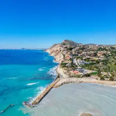 Vista aérea de la costa con casas blancas y el mar azul turquesa. La cala Baeza se extiende hacia el horizonte, con una bahía protegida por un rompeolas de rocas.