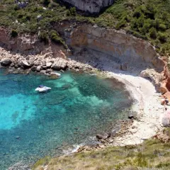 Aguas turquesas y una cala rocosa en Cala Llebeig, con un pequeño barco y casitas blancas en la orilla.