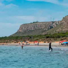 Gente disfruta del sol en la playa de la ermita santa pola, con aguas turquesas y un acantilado verde al fondo.