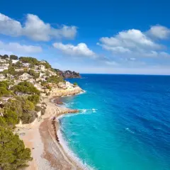 La **cala Baladrar** se asoma entre casas blancas y pinos verdes, con aguas turquesas y un mar azul intenso bajo un cielo nublado.