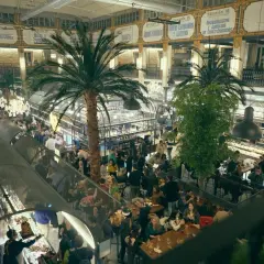 Un bullicioso mercado interior con palmeras, gente comiendo y comprando. El ambiente recuerda a un mercado de la imprenta, lleno de vida y actividad.