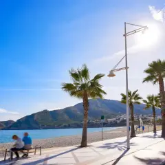 Pareja sentada en un banco en la playa cap blanc, con palmeras y montañas al fondo bajo un cielo azul brillante.