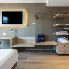 Habitación moderna con TV, escritorio, armario abierto con minibar y caja fuerte. Una bolsa azul con el logo de Senator Hotels descansa sobre un banco.