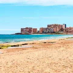 Gente pasea por la **playa de la Mata** con edificios residenciales al fondo y el mar azul.