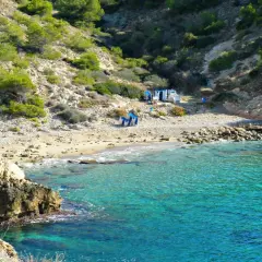 Agua turquesa en la cala Almadrava de Benidorm, con rocas y vegetación en la ladera. Hay algunas sillas y sombrillas en la playa.