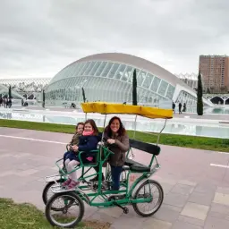 Familia disfrutando de un paseo en bicicleta en el **parque Gulliver**, con la Ciudad de las Artes y las Ciencias de Valencia de fondo.