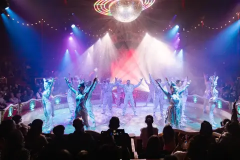 Artistas del circo Raluy en Alicante, bajo una bola de discoteca, con confeti cayendo y público aplaudiendo.