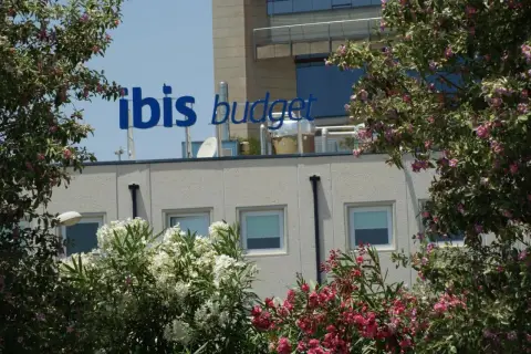 El cartel azul "ibis budget" destaca sobre el edificio del hotel Ibis Budget Alicante, rodeado de exuberante vegetación y flores rosadas y blancas.