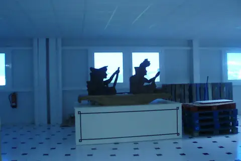 Siluetas de figuras en un interior, quizás una fábrica valor Alicante, con palets y un extintor.