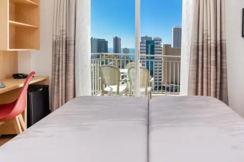 Habitación de hotel pueblo Benidorm con vistas al mar y rascacielos. Balcón con mesa y sillas. Escritorio con silla roja y minibar.