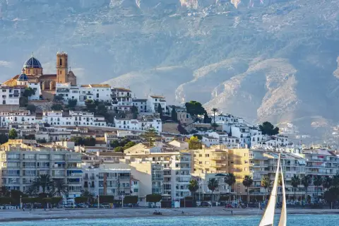 Vista del **castell de l'Olla** coronando la colina, con sus casas blancas y la iglesia de cúpula azul. Un velero navega frente a la costa.