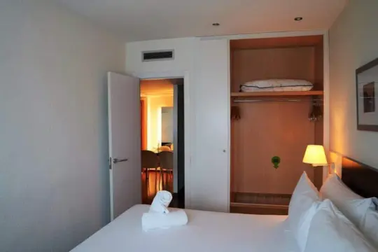 Habitación de hotel Port Feria Valencia. Cama blanca con toallas dobladas, armario abierto con perchas y una almohada. Puerta entreabierta a un comedor.