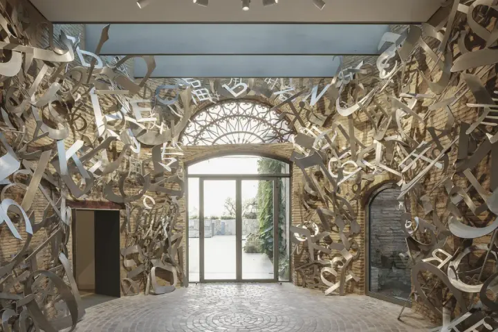 Instalación de arte abstracto con letras metálicas flotantes, evocando la obra de **hortensia herrero**, en un pasillo con paredes de ladrillo y suelo de adoquines.