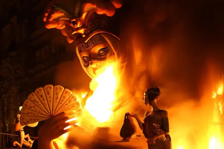 Figuras de las Fallas arden en llamas, evocando el origen de esta tradición. Una mano sostiene un abanico, mientras otra figura observa el fuego.