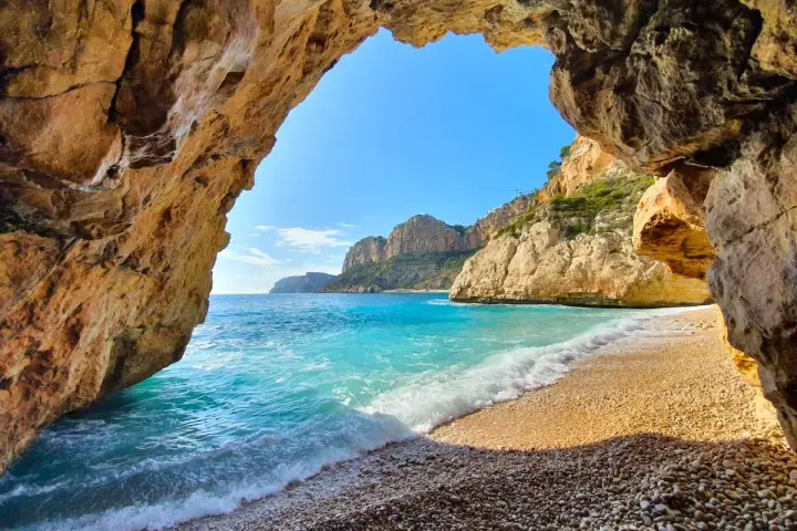 Vista desde una cueva hacia la cala dels testos, con aguas turquesas, arena de guijarros y acantilados rocosos.