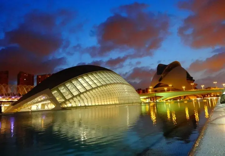La **Ciudad de las Artes y las Ciencias** de Valencia, con sus icónicos edificios futuristas, se refleja en el agua al anochecer, bajo un cielo dramático.
