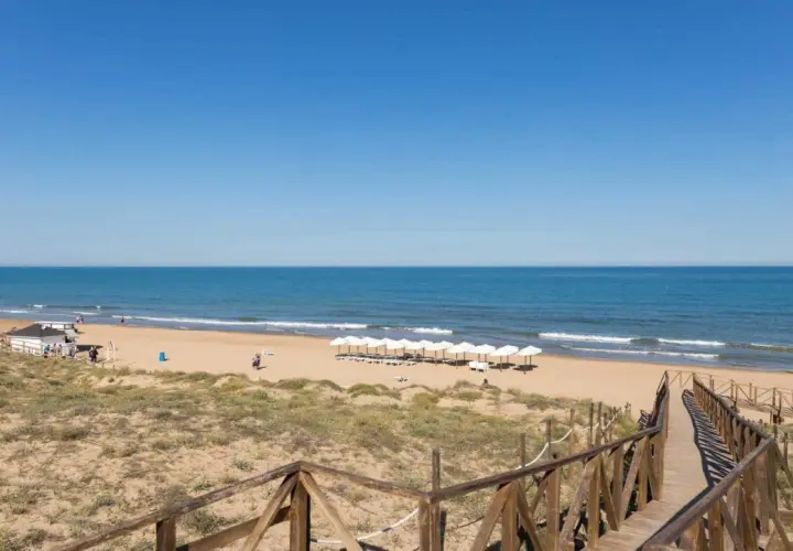 Pasarela de madera hacia la playa de Miramar, con sombrillas blancas alineadas y el mar azul bajo un cielo despejado.